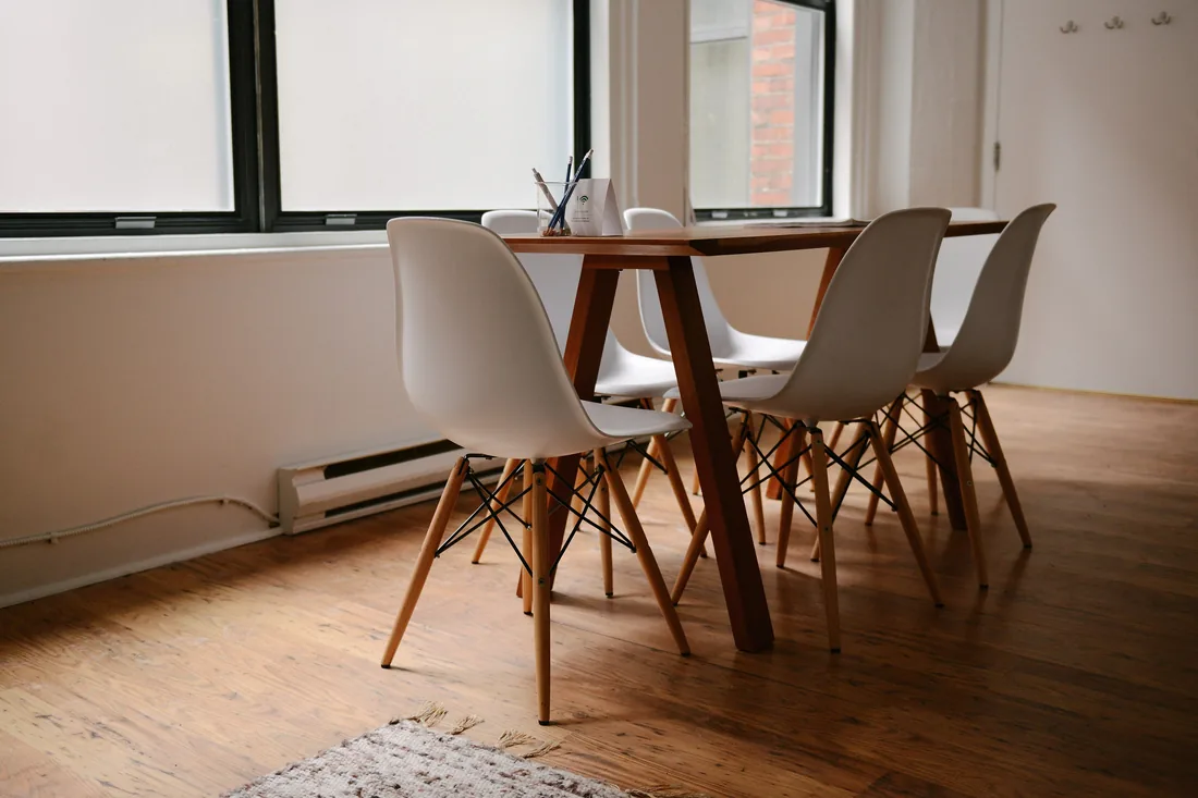An empty room with a wooden table, white chairs and daylight through tall windows